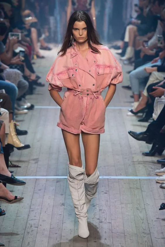 На девушке свободный нежно-розовый комбинезон с карманами, заклепками и шнуровкой на талии и белые высокие широкие сапоги с заклепками.